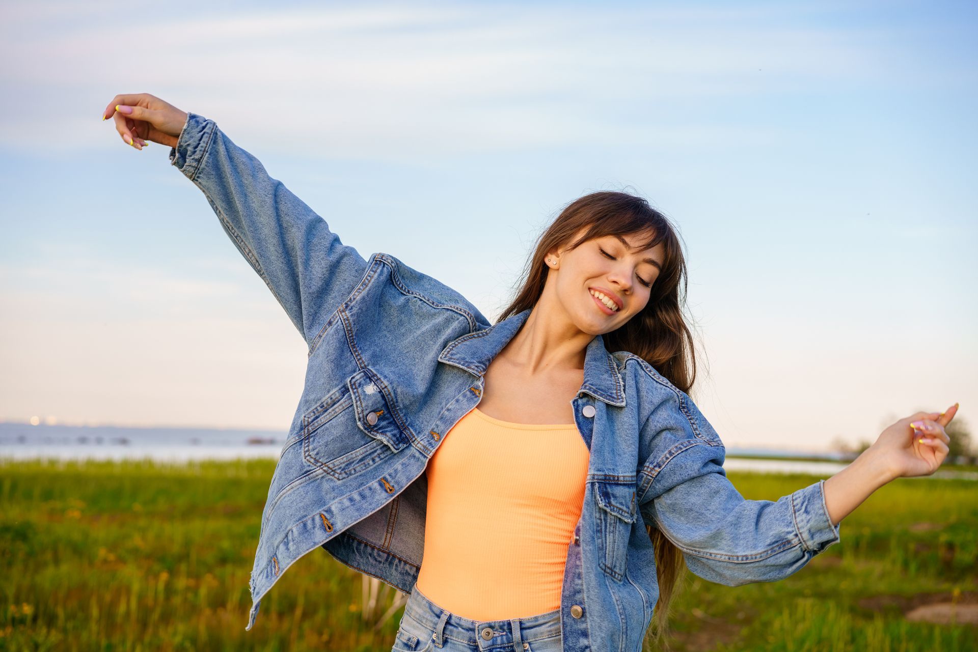 A woman in a denim jacket and orange tank top is standing in a field with her arms outstretched.