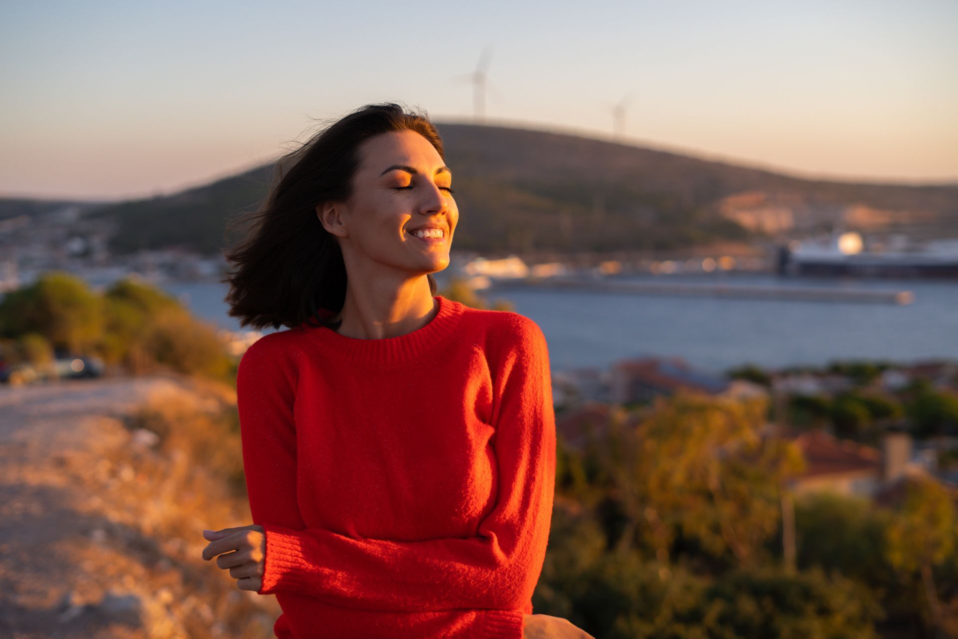 A woman in a red sweater is standing on top of a hill overlooking the ocean.