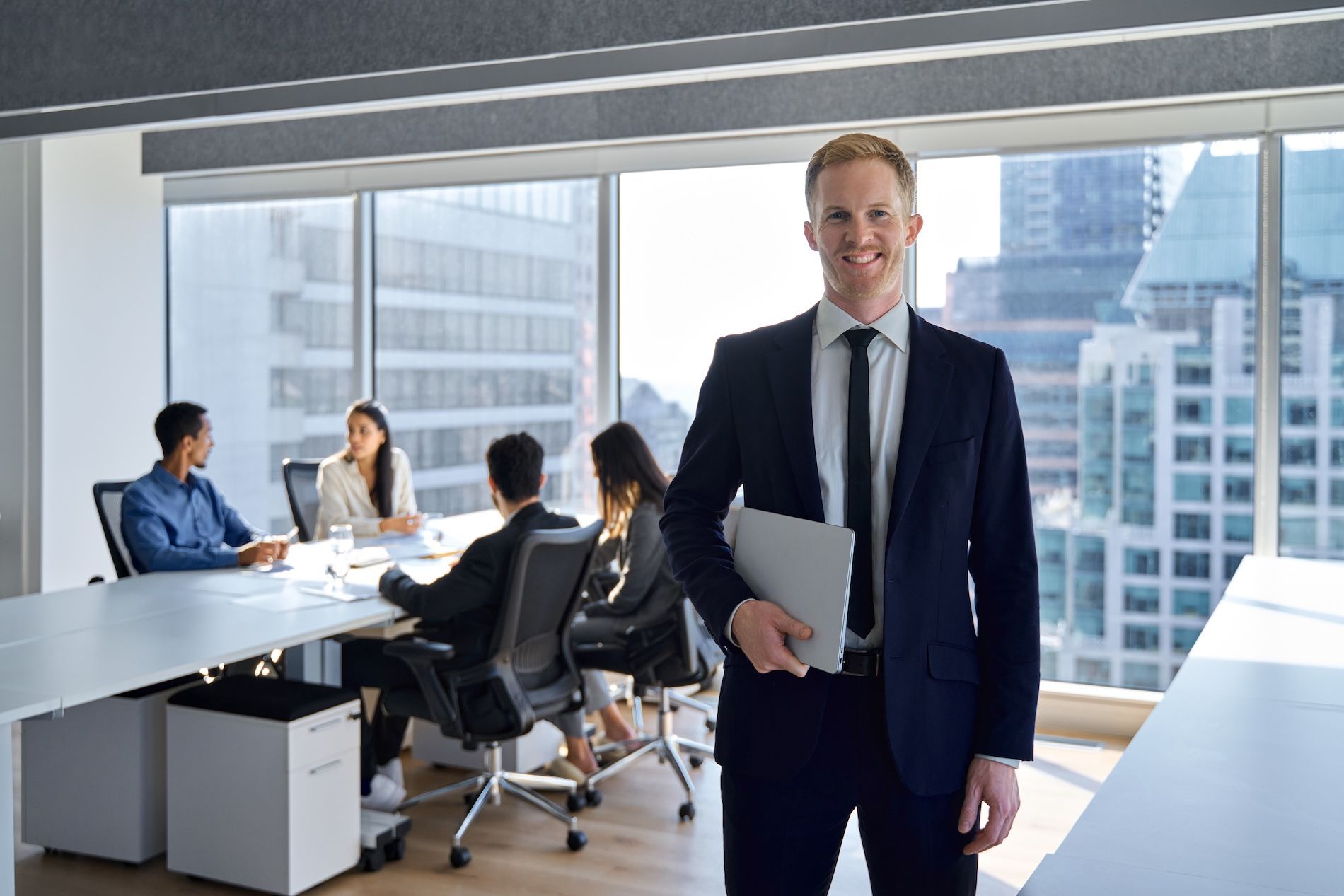 Man in suit holding tablet, smiles in office with meeting in progress.