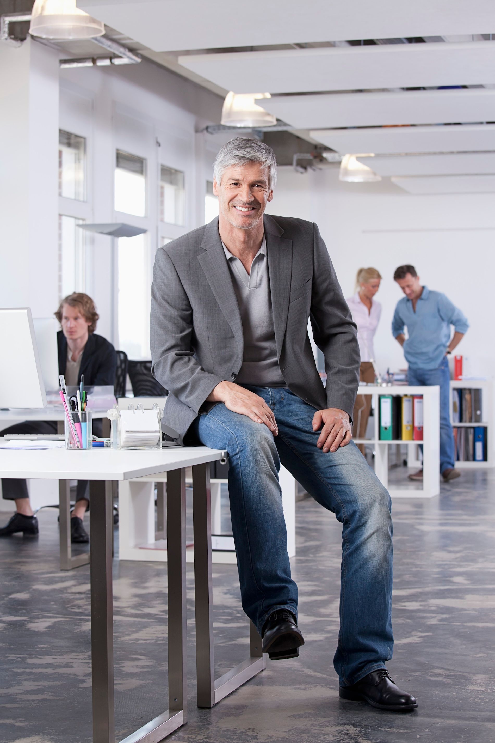 Man in gray blazer and jeans, sitting on a desk in a bright office; colleagues in background.