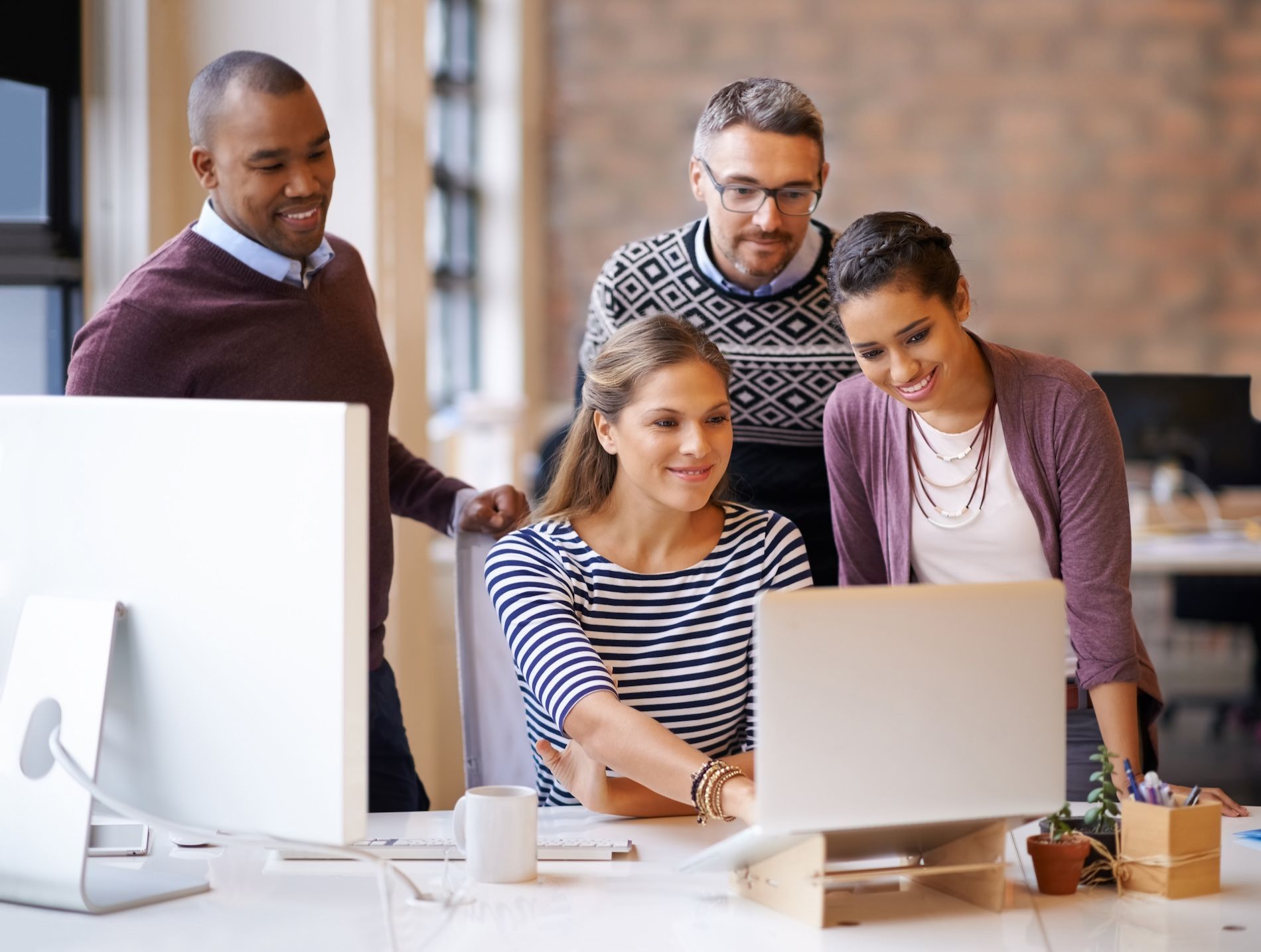 Office colleagues gathered around a laptop, smiling and collaborating at a desk.
