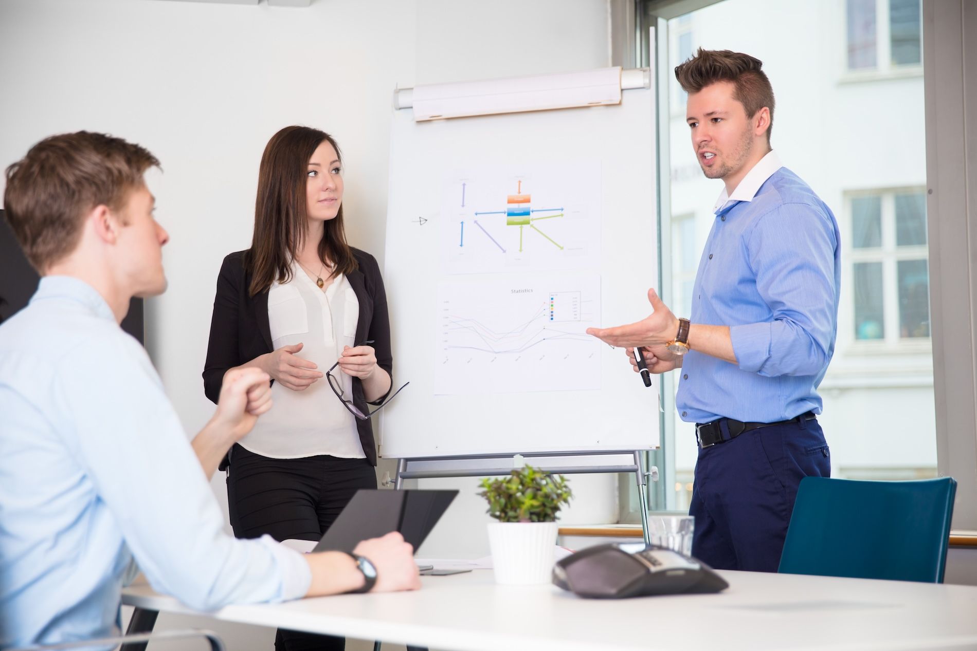 Three business people in a meeting. Man presenting at whiteboard, woman holding glasses, and man at the table looking on.