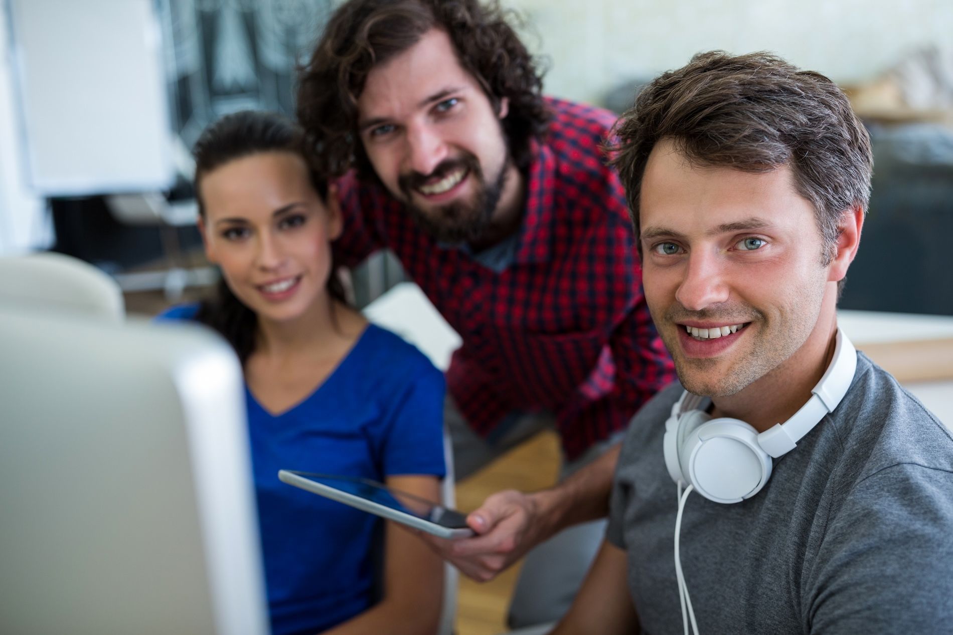 Three smiling colleagues looking at a computer screen. One holds a tablet; headphones on the man in front.