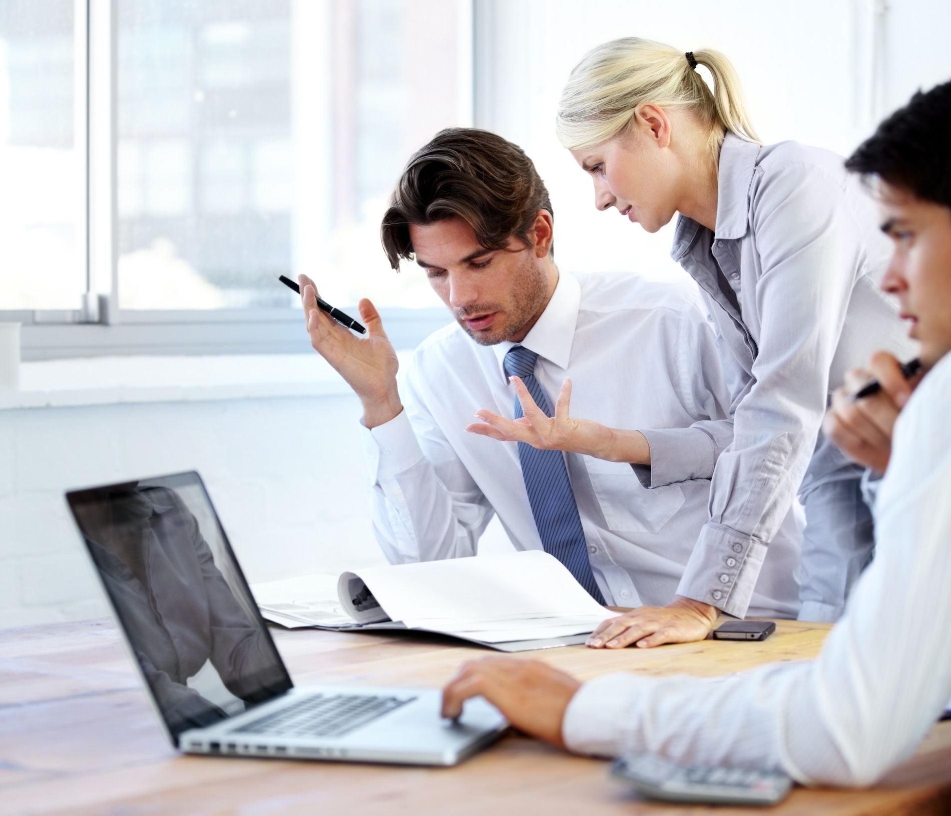 Three business colleagues reviewing documents at a table, near a laptop and window.
