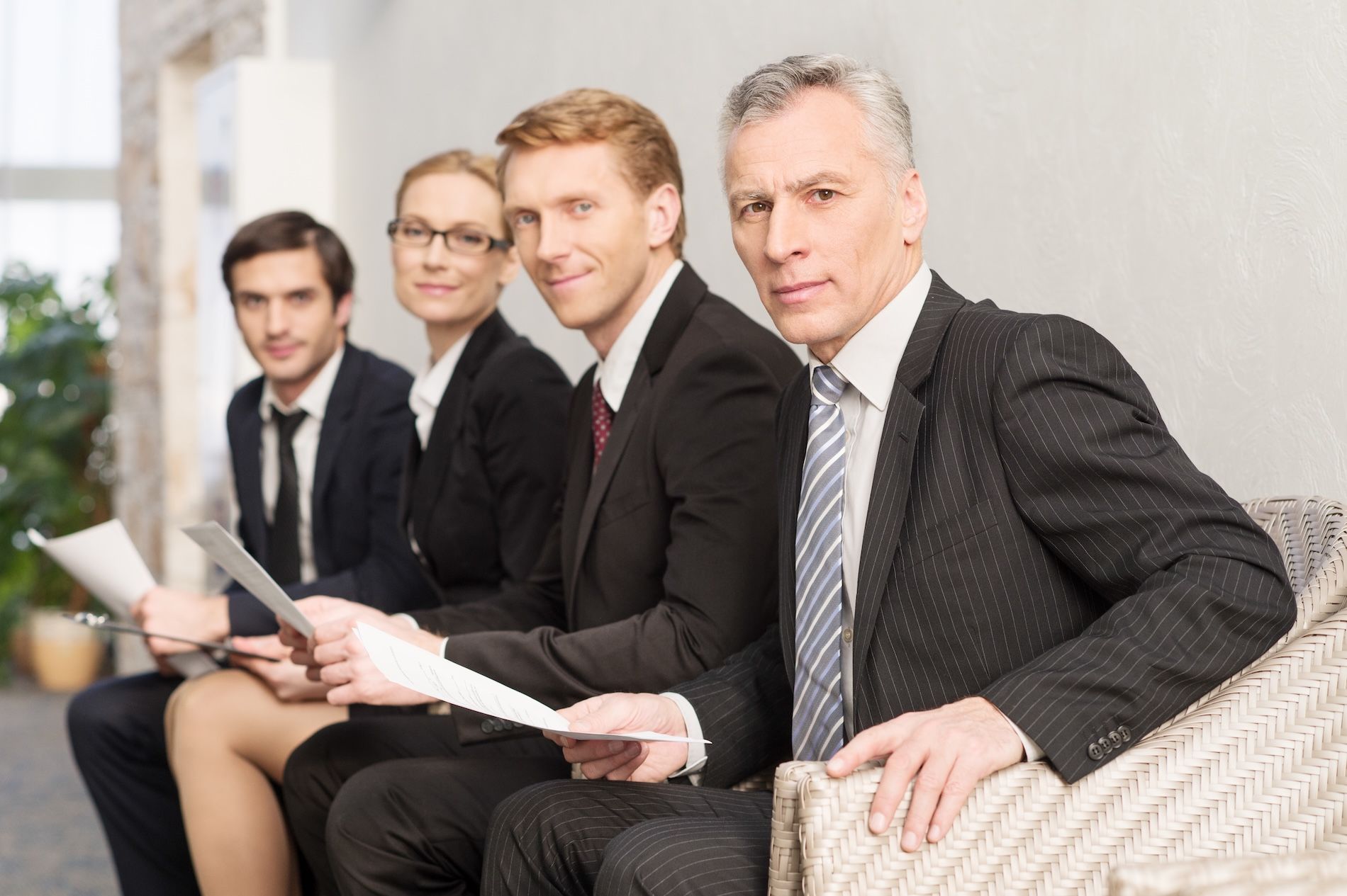 Four formally dressed people seated, holding papers, looking at the viewer.