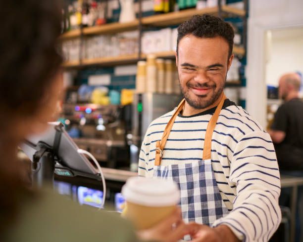 Man with Down syndrome smiles, handing a coffee cup to a customer at a cafe counter.
