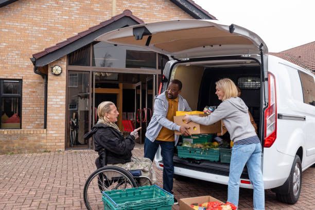 People loading boxes of food from a van; a person in a wheelchair watches at the building entrance.