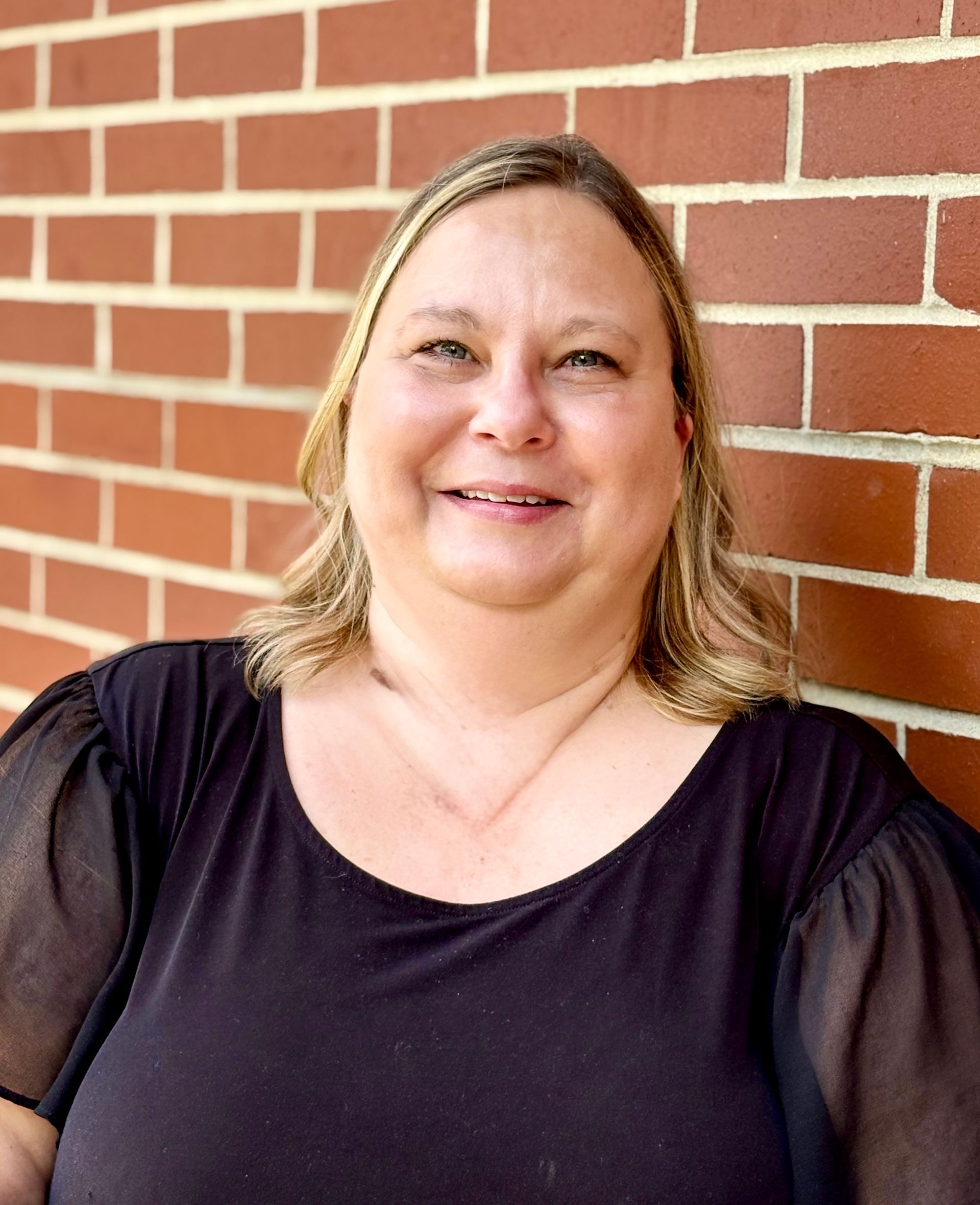 Woman with blonde hair smiles, wearing a black top, stands in front of a red brick wall.