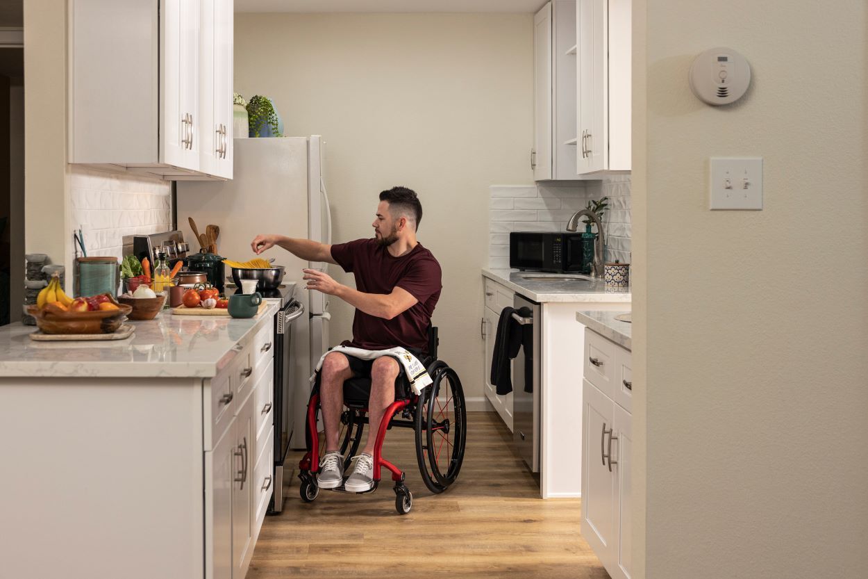 Man in wheelchair cooking in a white kitchen. He reaches for something, preparing food.