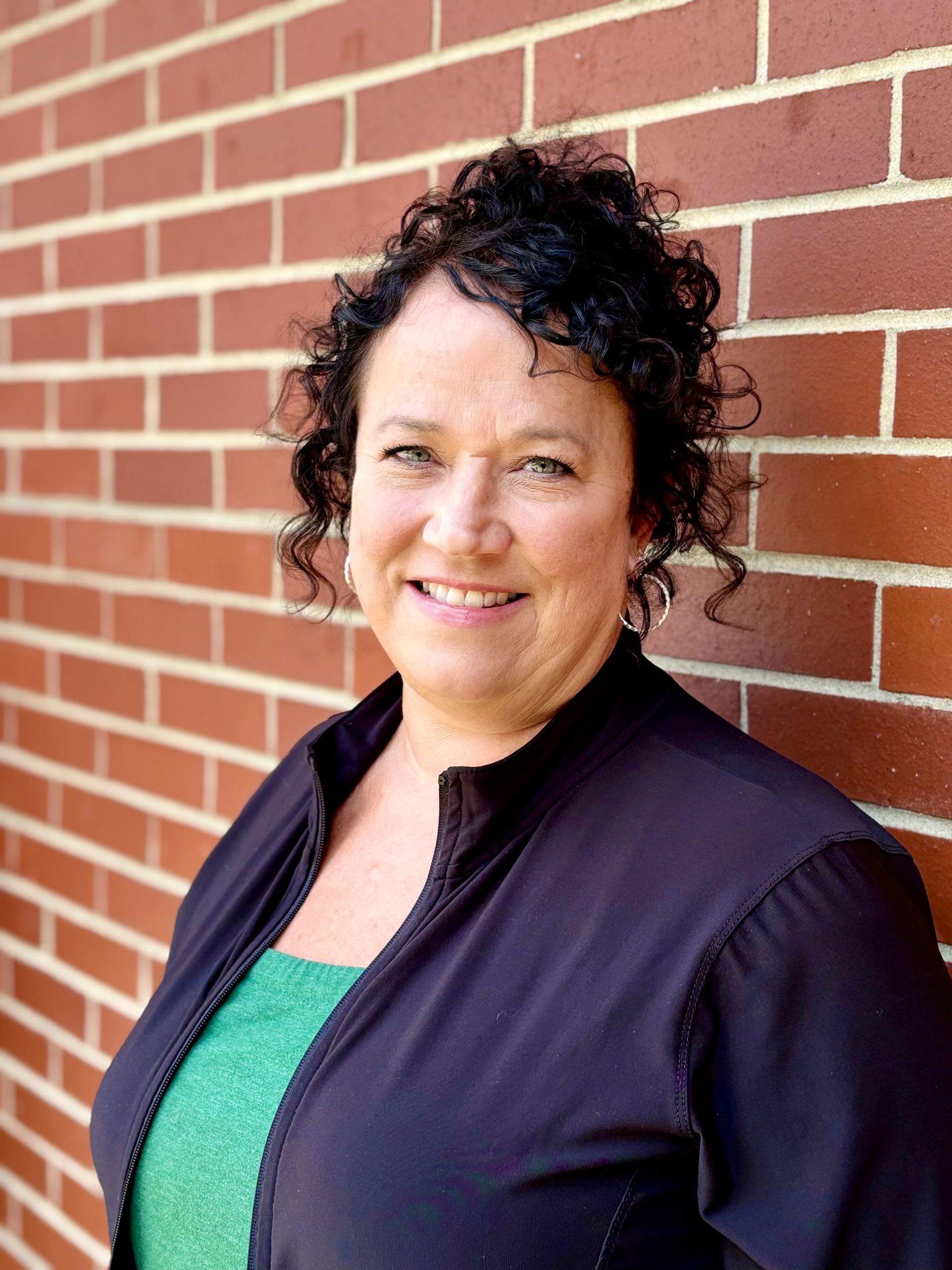 Woman with curly dark hair smiles in front of a brick wall. She wears a black jacket over a green top.