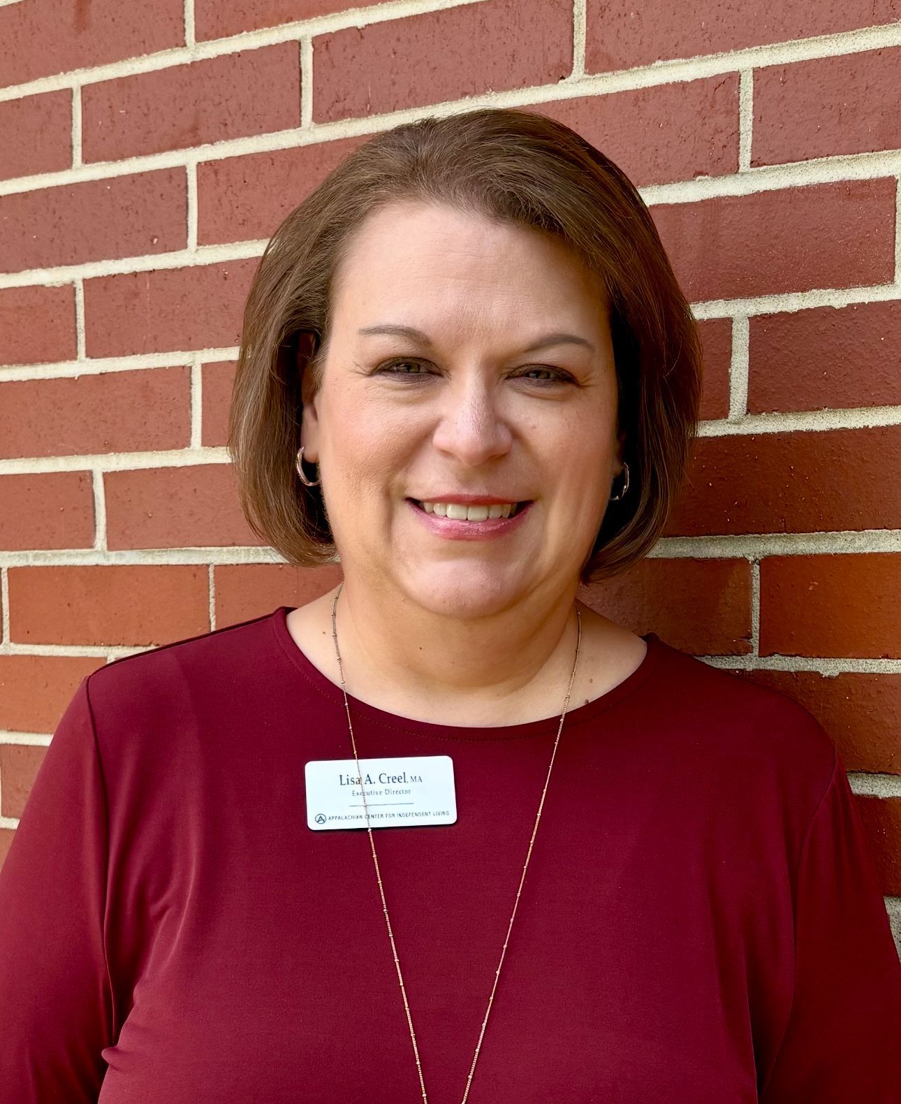 Woman with short brown hair, smiling, in front of a brick wall, wearing a maroon shirt and name tag.