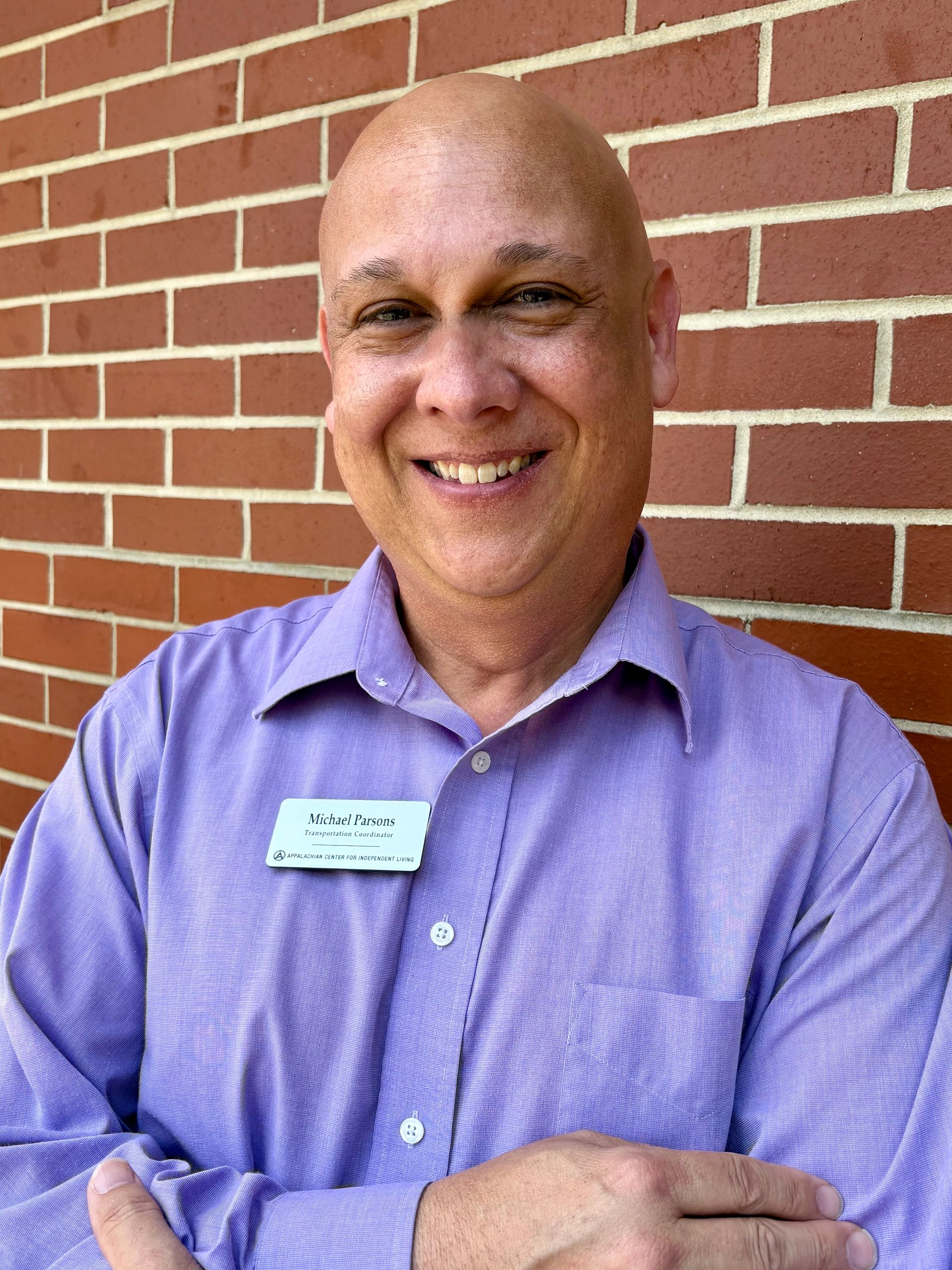 Bald man wearing a purple shirt smiles in front of a brick wall, a name tag on his shirt.