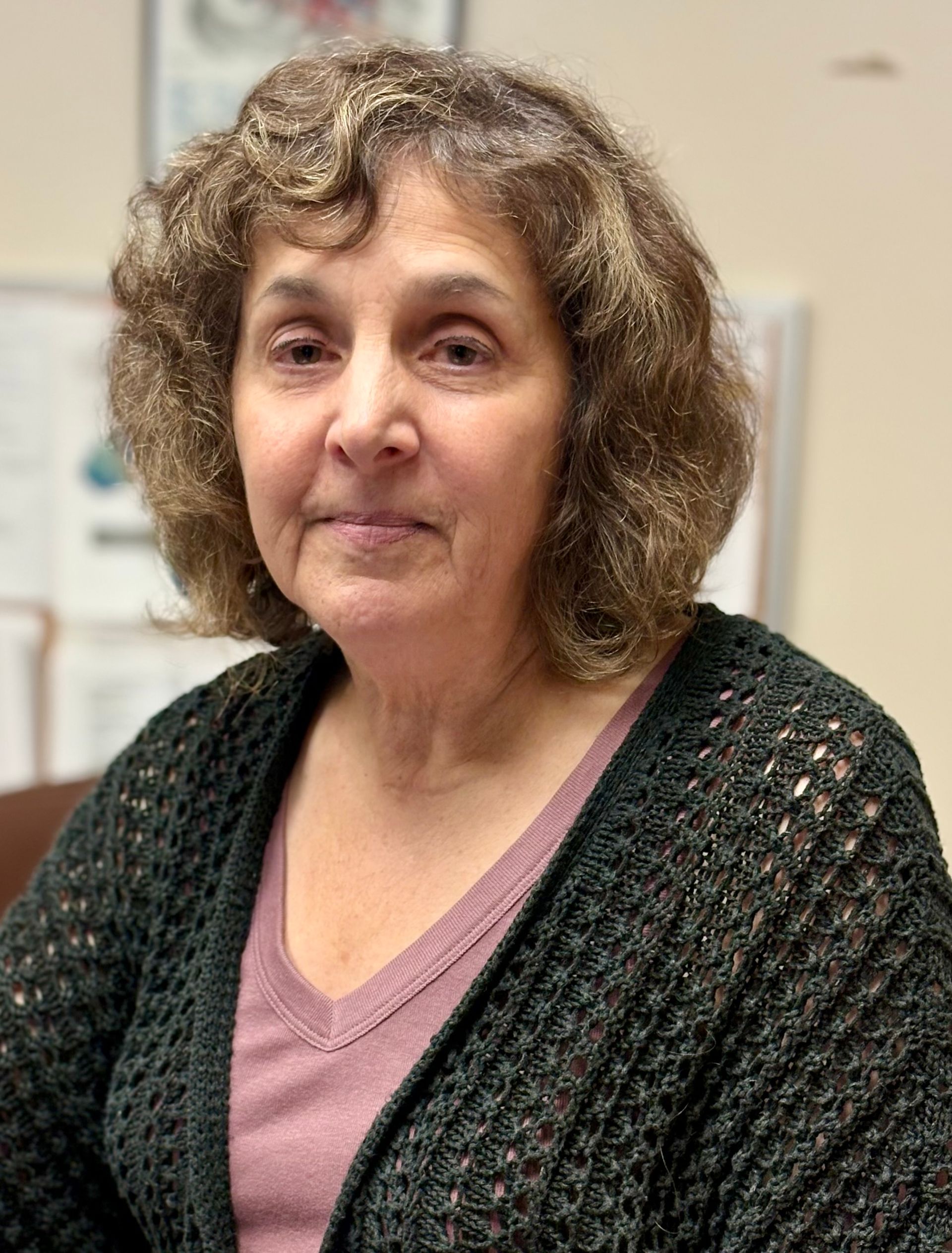 Woman with curly brown hair, wearing a dark green cardigan over a pink shirt, smiling slightly.
