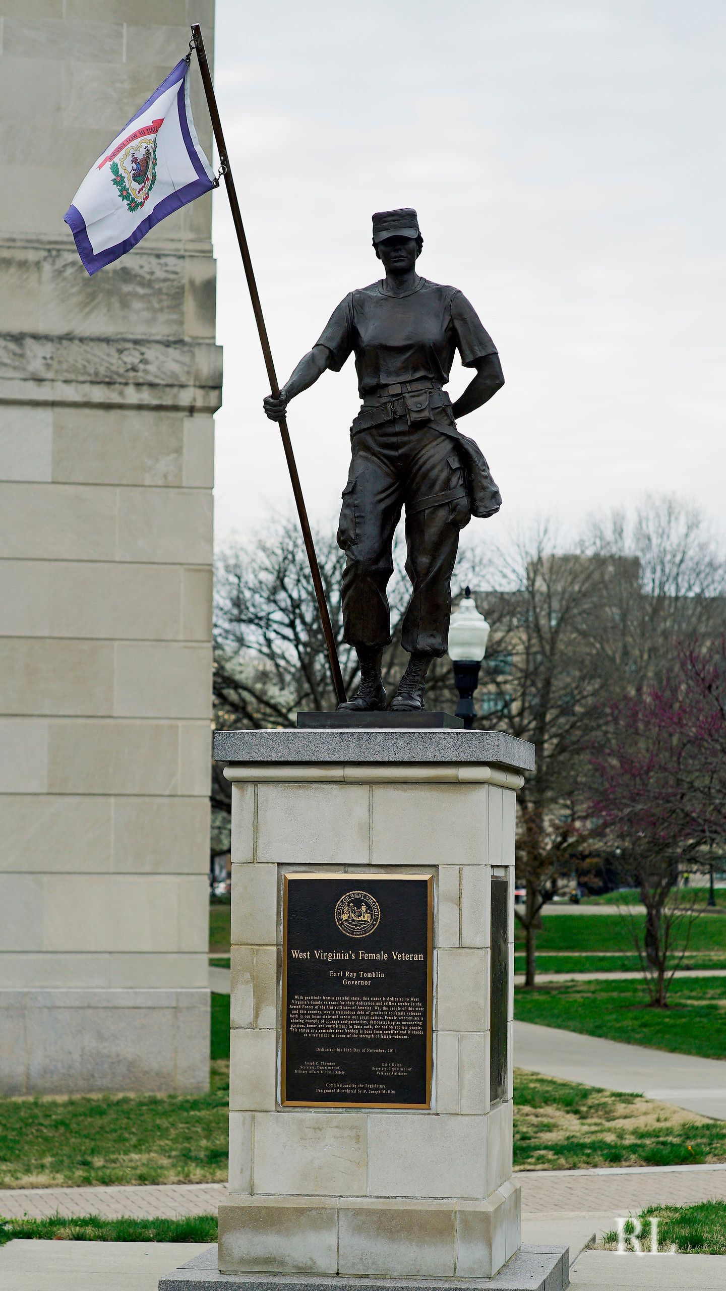 A bronze statue of a soldier holding a flag on a stone pedestal in a park-like setting.