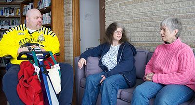 Man in wheelchair talks to two women on a couch in a room with a brick wall and bookshelf.