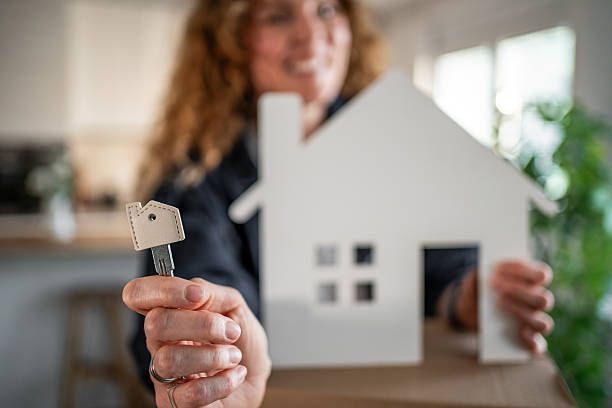 Woman holding a key and a white house cutout; smiling, indoors.