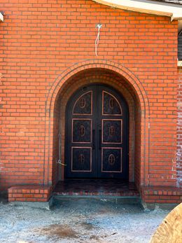 A dark metal double door with decorative panels and an arched top, set into an unfinished red brick wall.