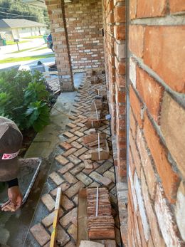 A person installing a herringbone brick pattern on a walkway against a red brick house wall.