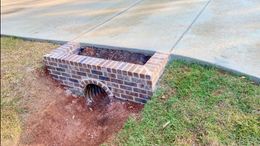 A brick-encased culvert headwall positioned alongside a concrete driveway in a grassy yard.