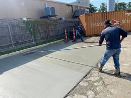 A worker uses a long-handled tool to smooth wet concrete in an outdoor work area next to a shipping container.