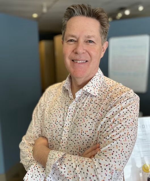 A man in a white shirt with polka dots is smiling with his arms crossed