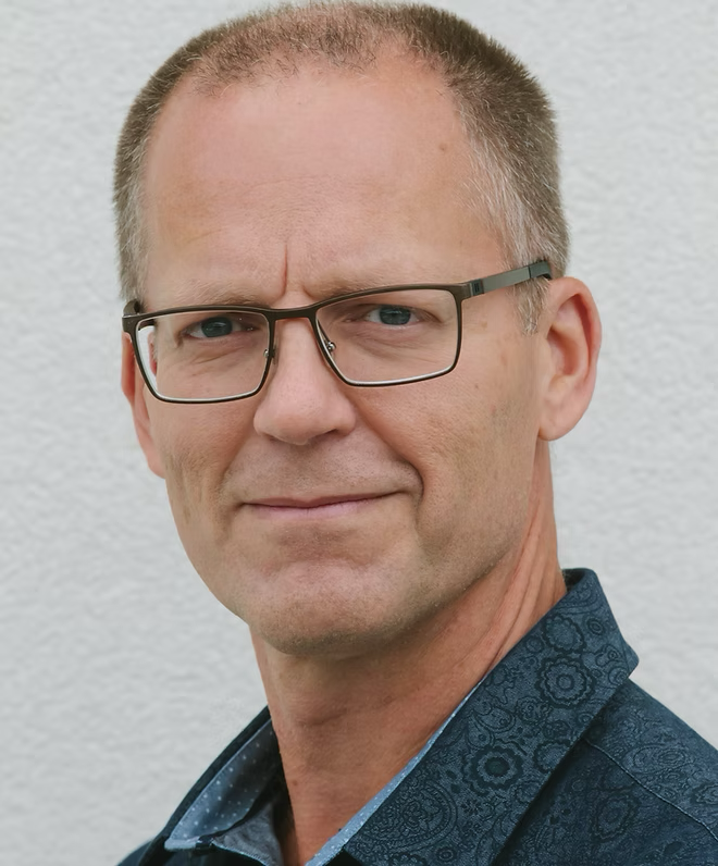 A man wearing glasses and a blue shirt is looking at the camera.