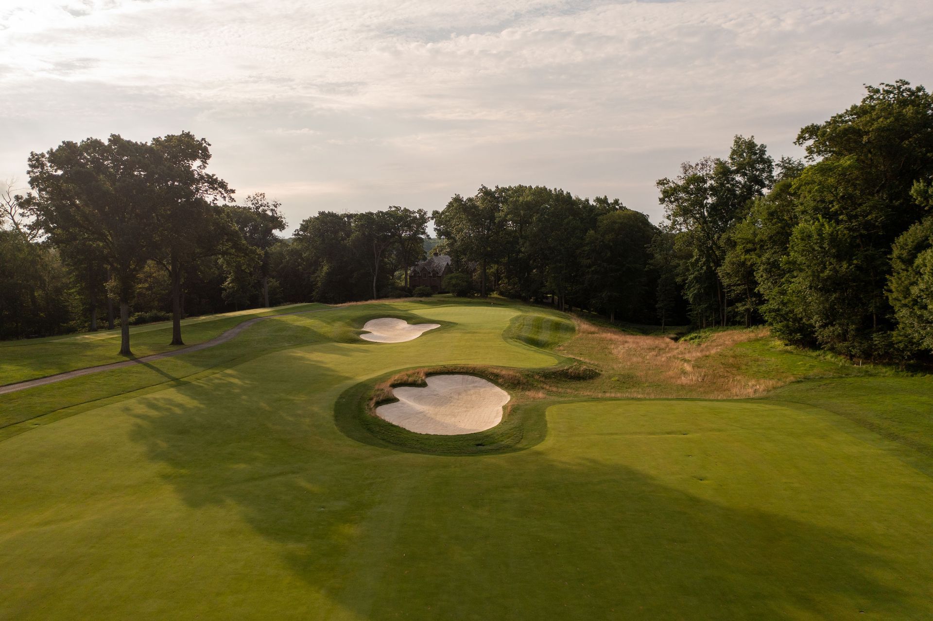Green golf course with two sand traps, trees, and cloudy sky.