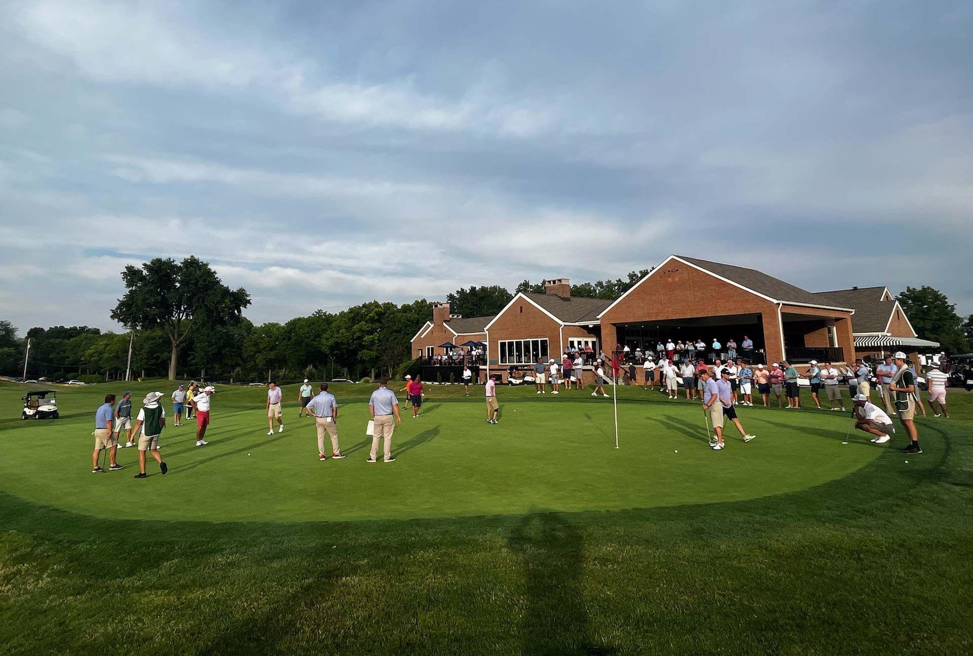 Golfers on a putting green in front of a clubhouse with spectators. Overcast sky.