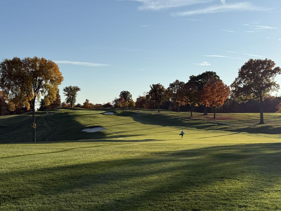 Golf course on a sunny day, featuring green grass, autumn trees, and a blue sky.