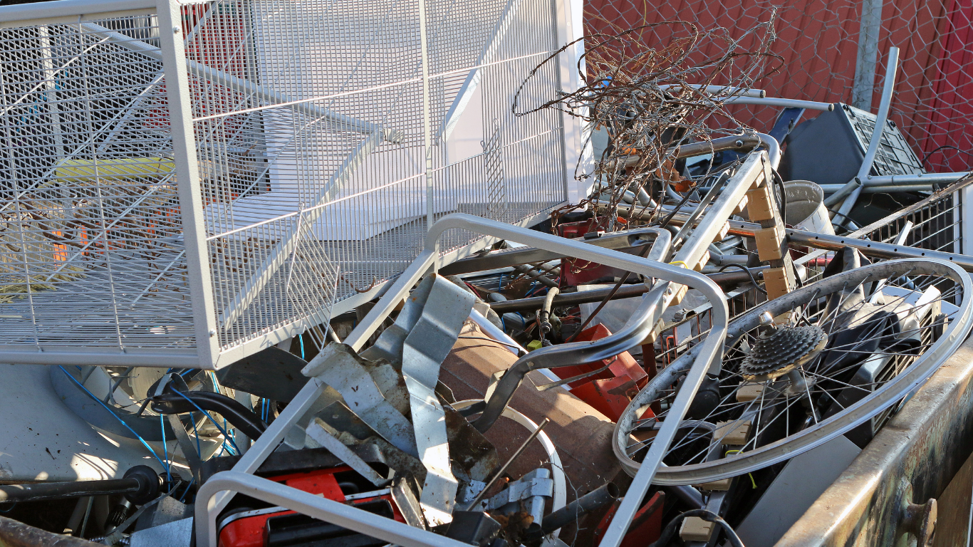 A pile of scrap metal including a shopping cart, bicycle parts, and miscellaneous items, outdoors.