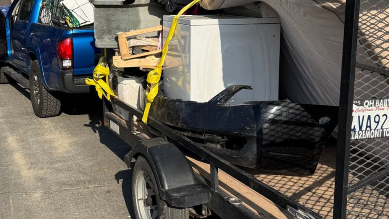 A blue pickup truck towing a trailer loaded with junk, including a washing machine and bumper.