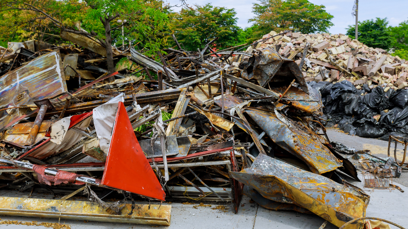 Pile of charred debris, including metal and wood, likely from a fire. Orange warning sign visible.
