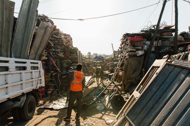 A worker in an orange vest stands between piles of scrap. A truck is on the left.