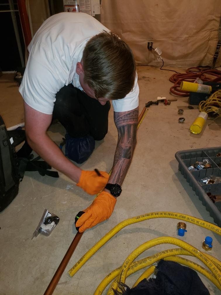Plumber kneeling, working on copper pipe. Wearing gloves, marking pipe. Tools and materials on floor.