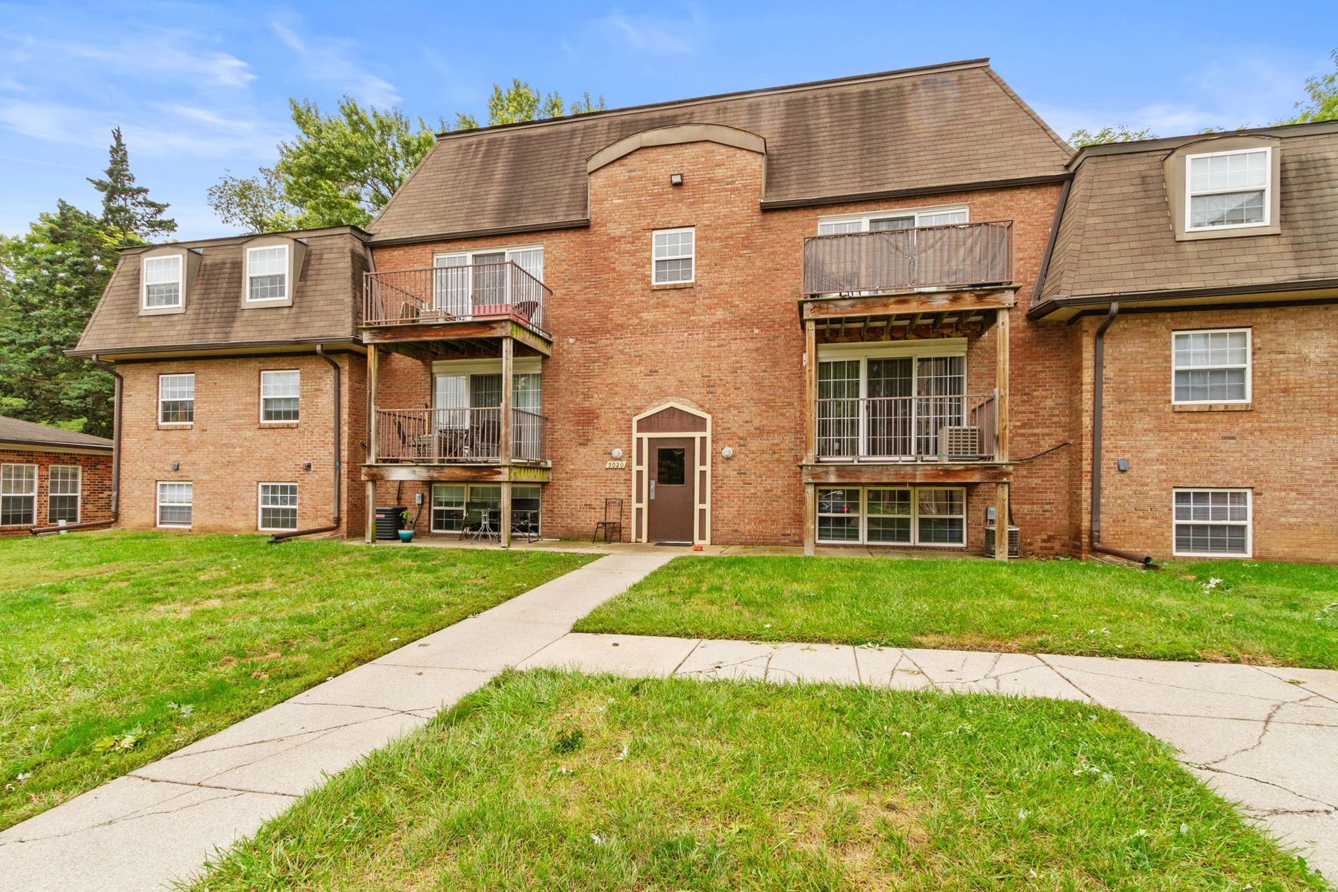 Red brick apartment building with balconies, a walkway, and green grass.