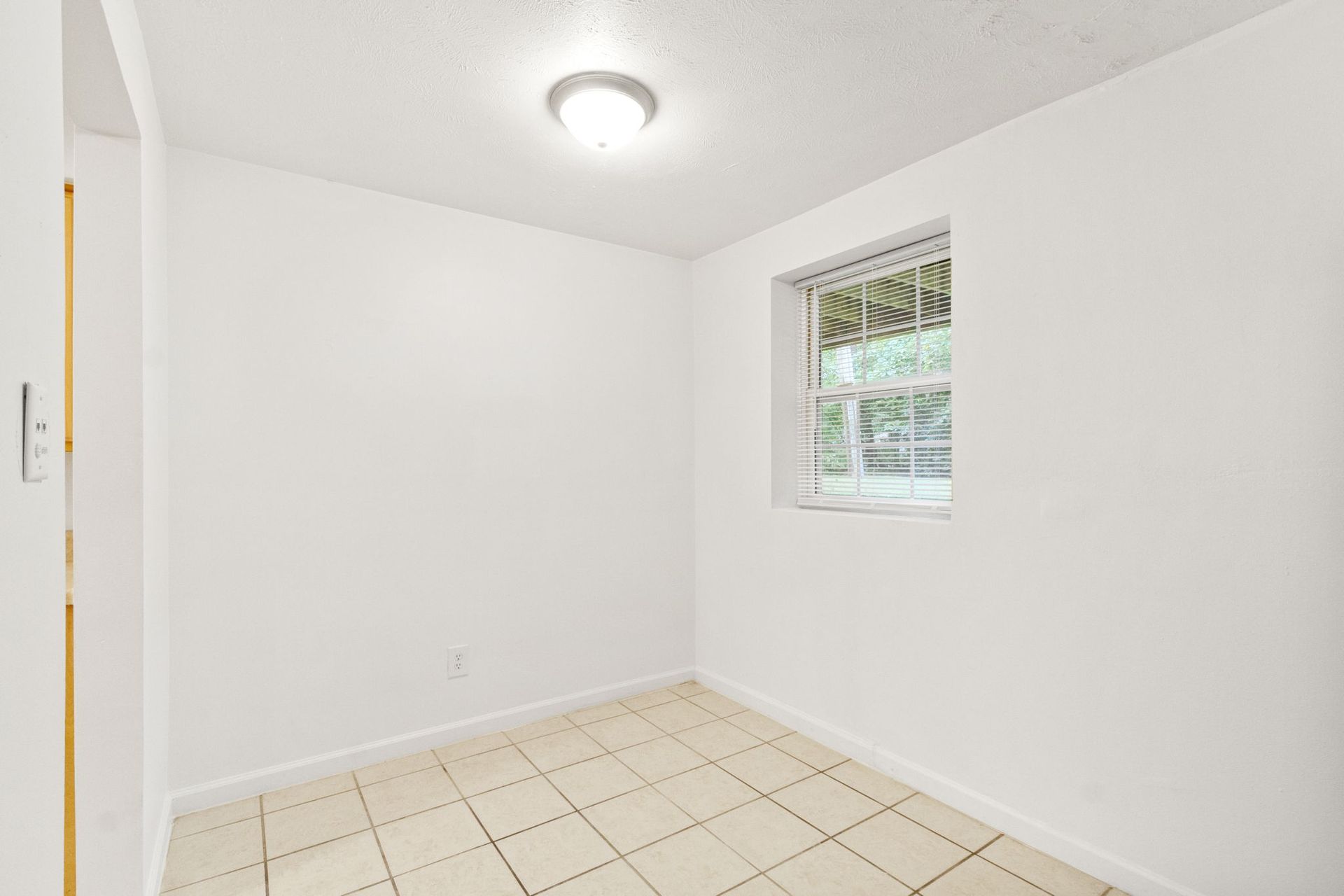 Empty room with white walls, tile floor, window with blinds, and a ceiling light.