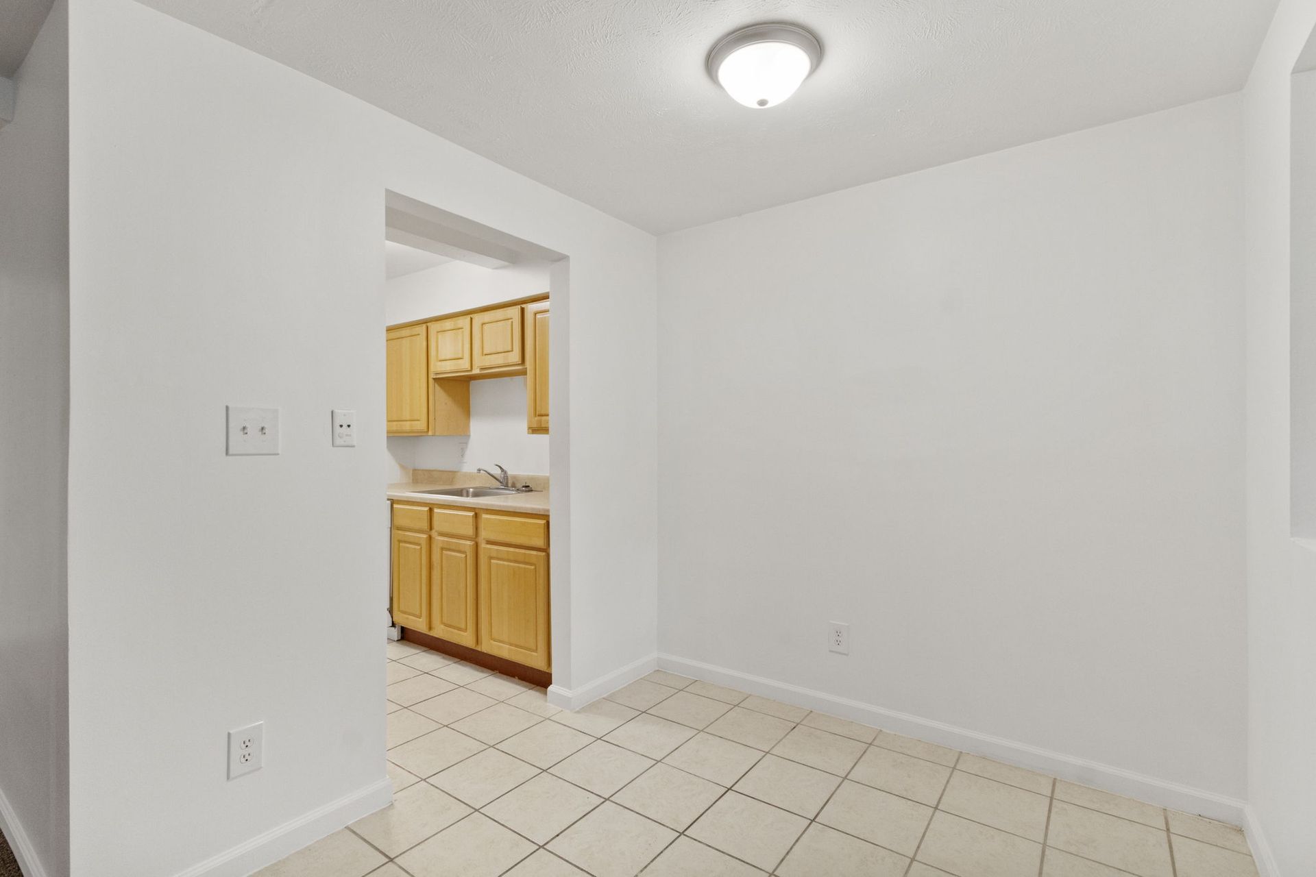 Empty dining room with kitchen visible through doorway. Light walls, beige tile floor, and a ceiling light.