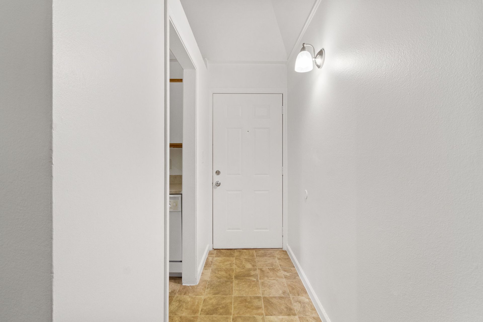 Hallway with white walls, brown tile floor, and white door at the end. A light fixture is mounted on the right wall.