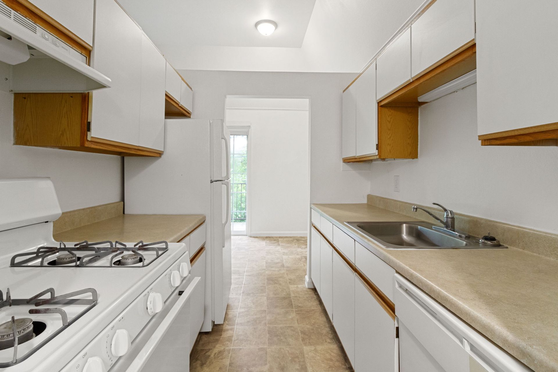 A long, narrow kitchen with white cabinets, appliances, and beige countertops and floor. Doorway at end.
