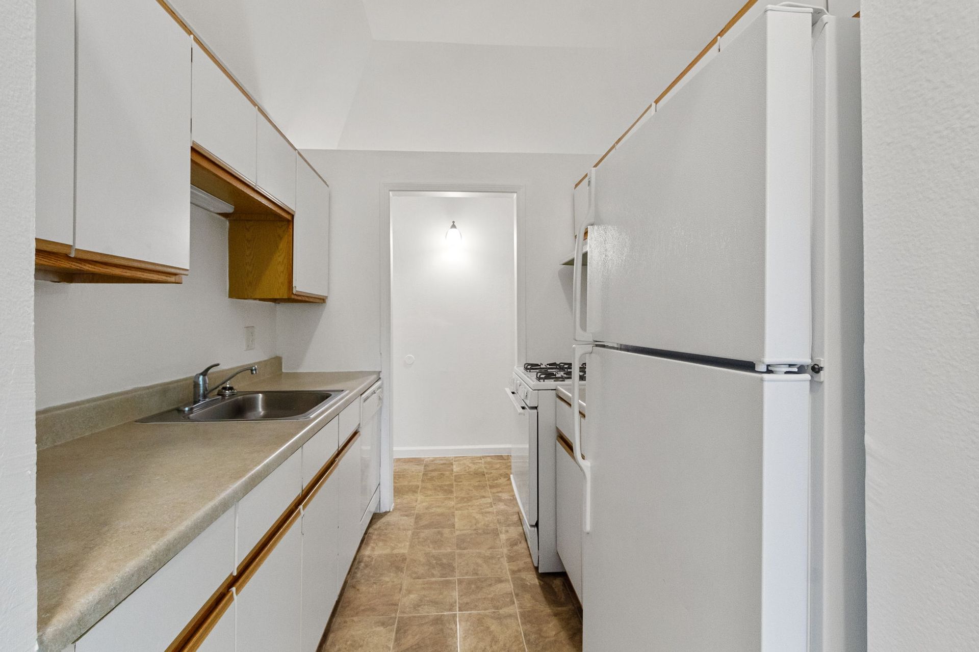 A narrow, white kitchen with a refrigerator, stove, sink, and cabinets.
