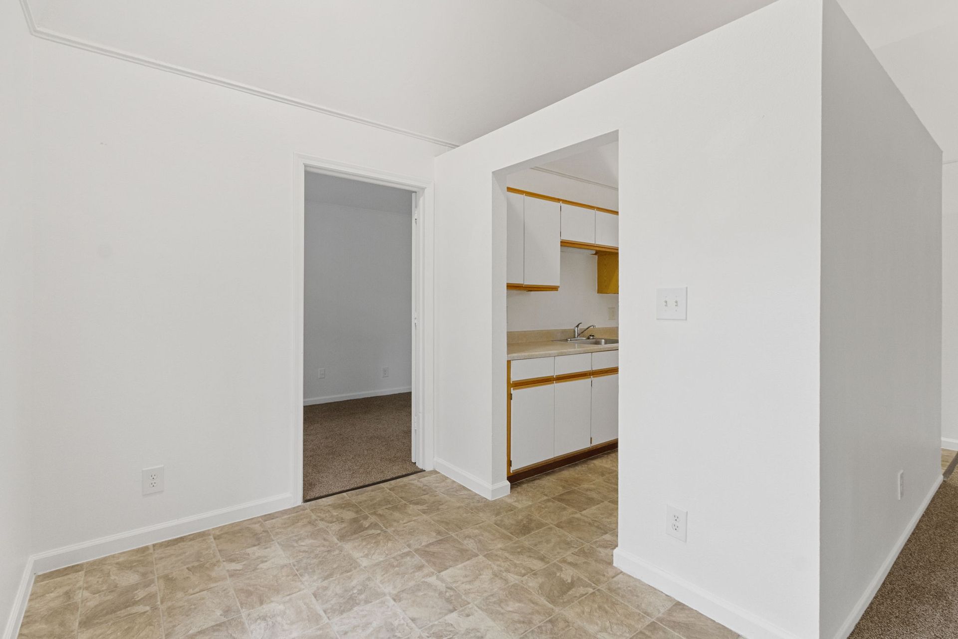 A white-walled interior with doorway to a kitchen and another room, beige flooring and carpet.