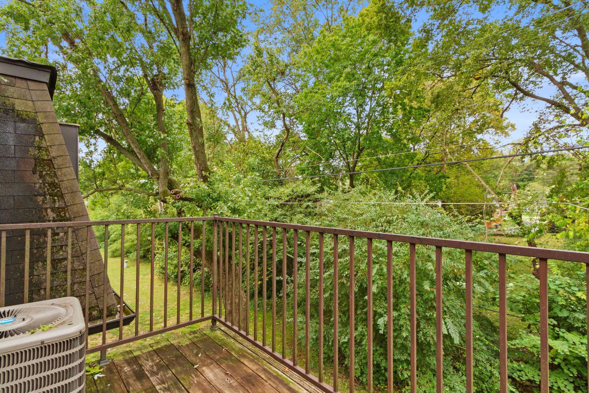 Balcony overlooking lush green trees, with a brown railing and air conditioner.
