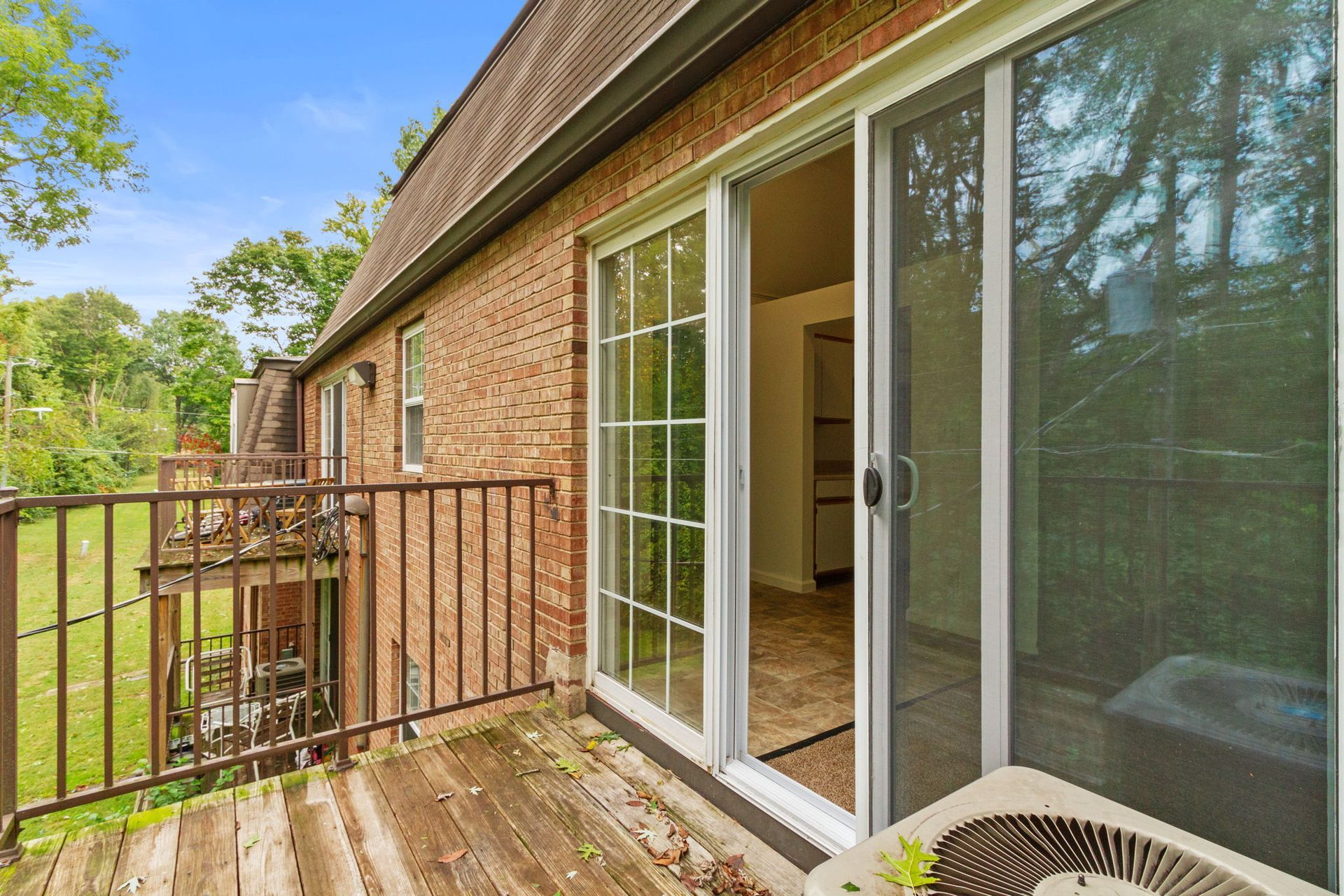 Balcony with sliding glass doors opening into a brick building, overlooking a wooded area on a sunny day.