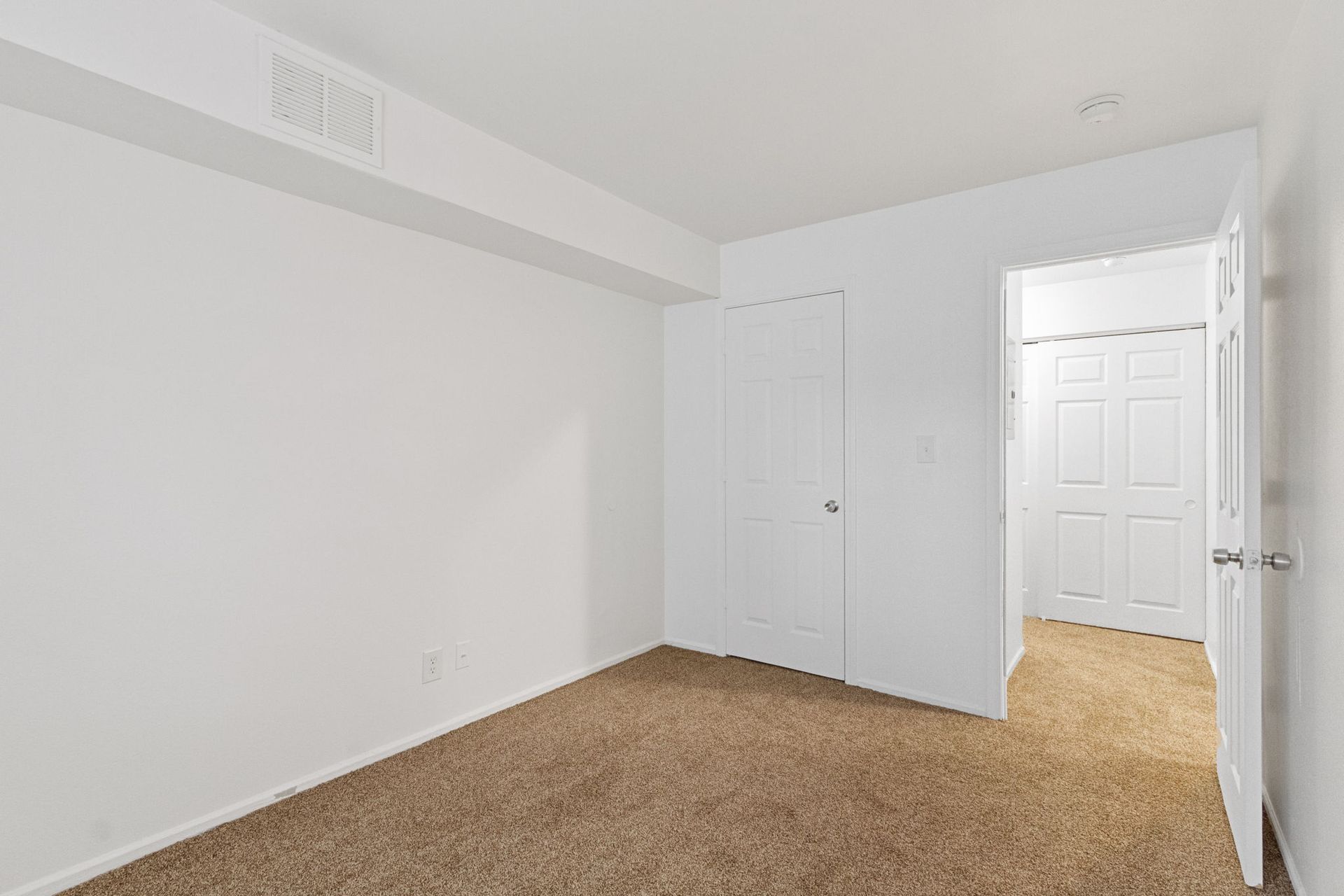 Empty room with brown carpet, white walls, and a closed door. Another door is visible in the opening.
