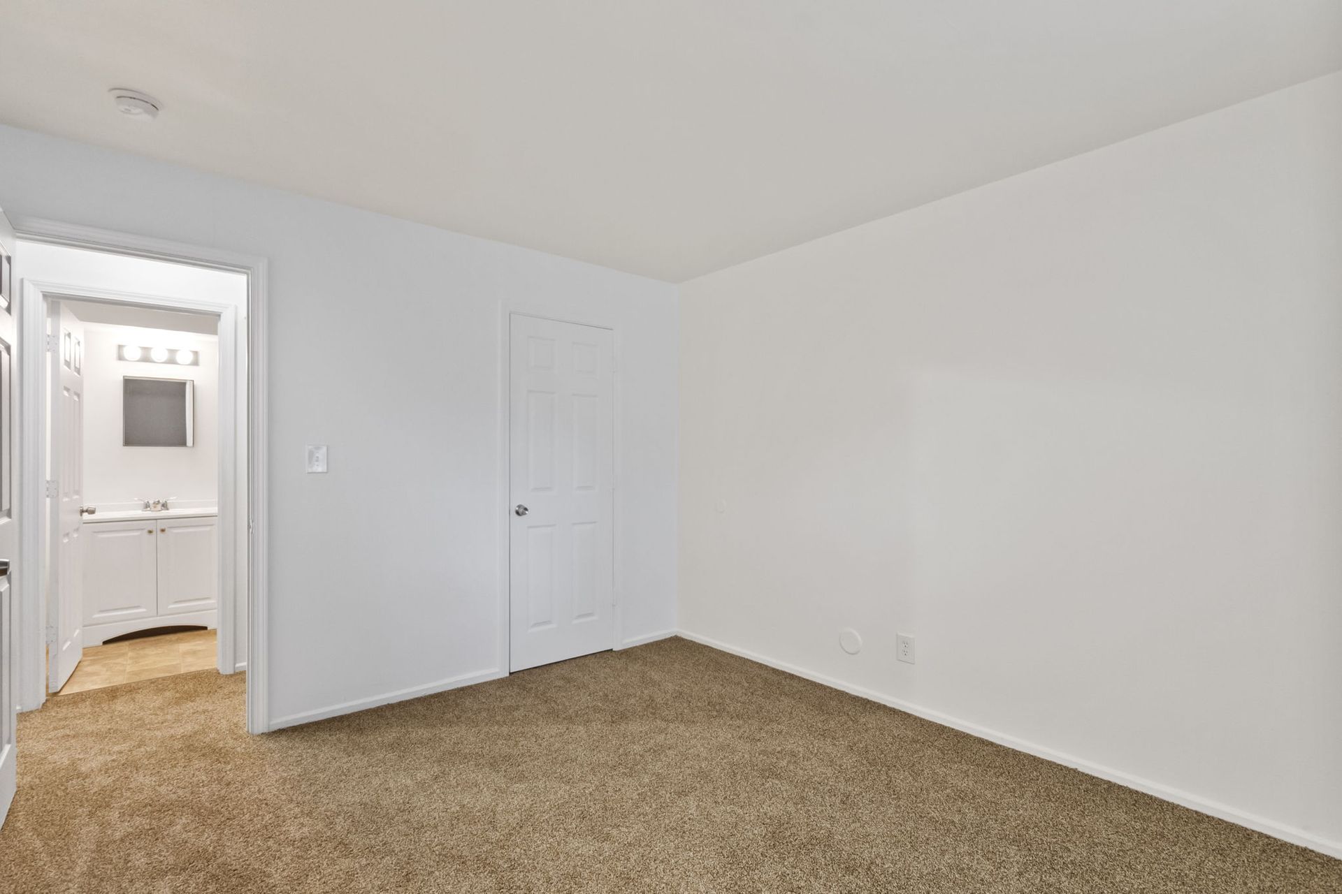 Empty bedroom with white walls, brown carpet, and a door to a bathroom.
