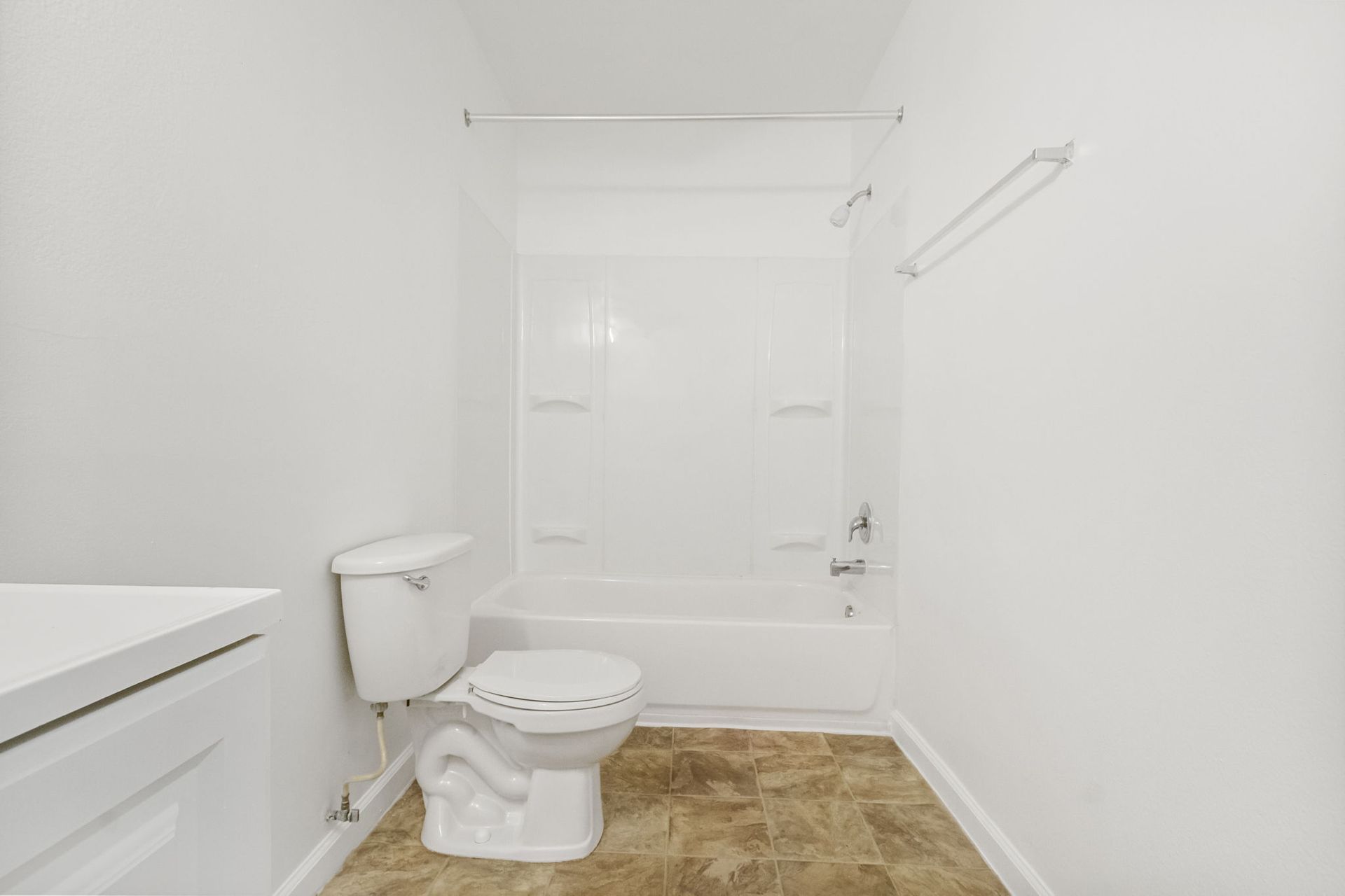A small, white bathroom with a toilet, bathtub, and a vanity. Brown flooring.