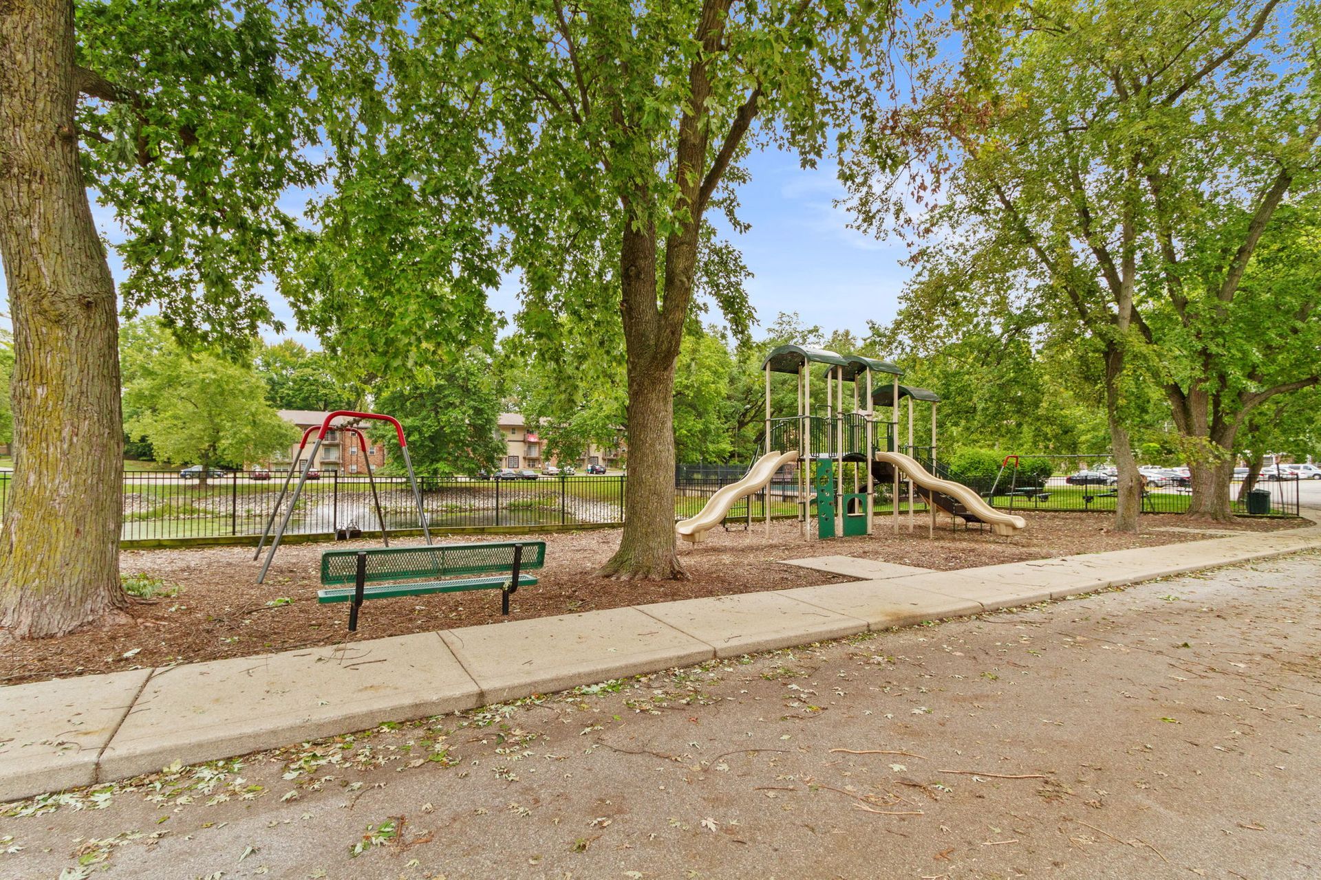 Playground with swings, slides, and bench, surrounded by trees and a fence.