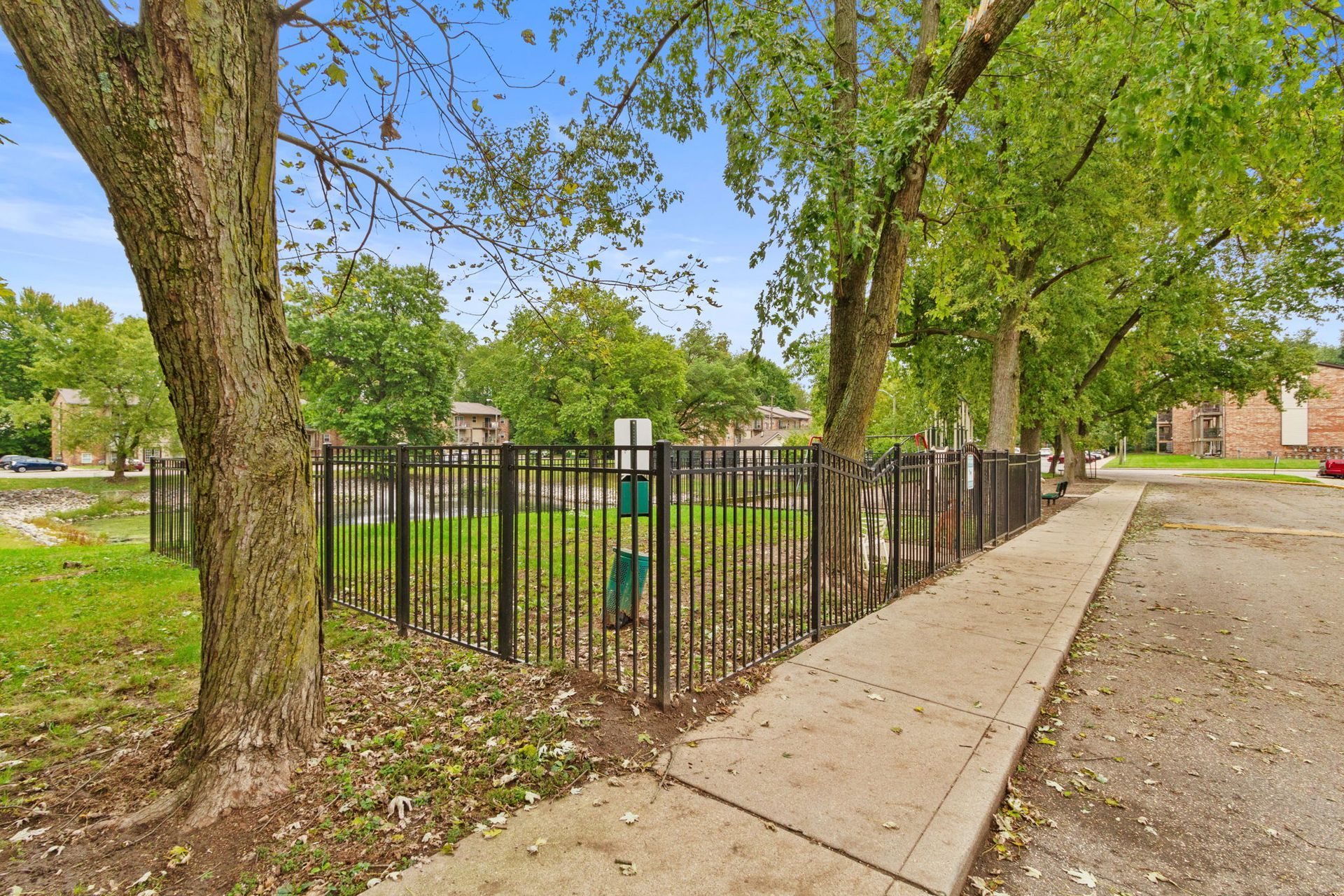 Fenced green space with trees and a sidewalk, possibly a dog park, in an outdoor residential area.