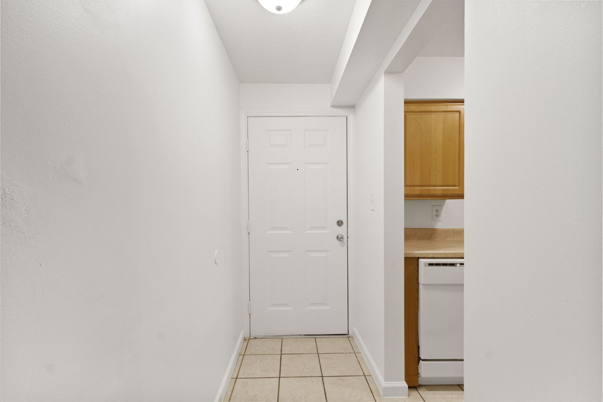 White hallway with a door at the end, leading to a kitchen with wood cabinets and a white dishwasher.