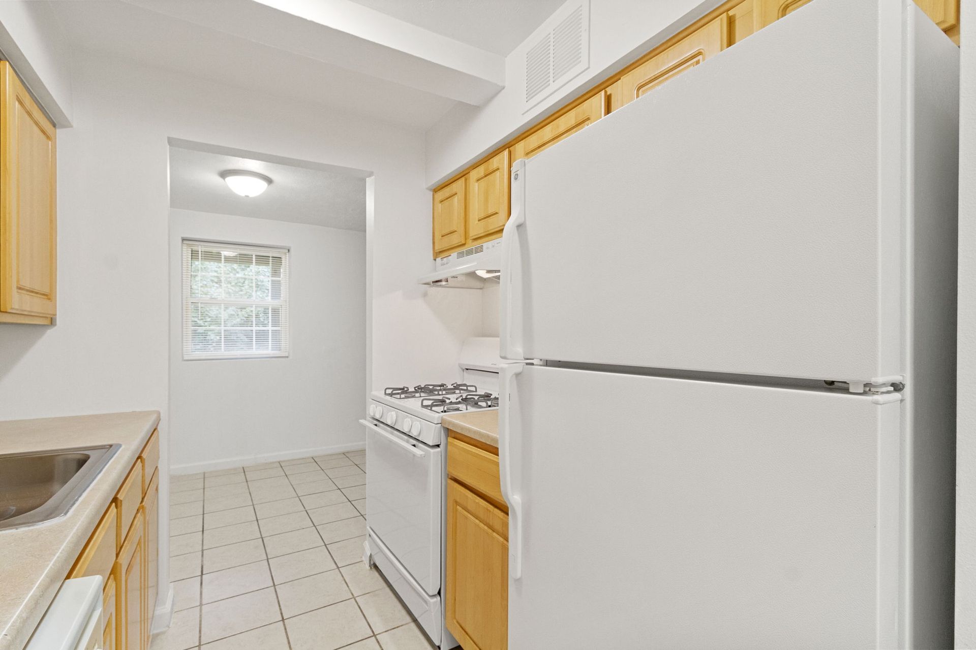 Kitchen with white appliances, light wood cabinets, and tile floor. An adjoining room has a window.